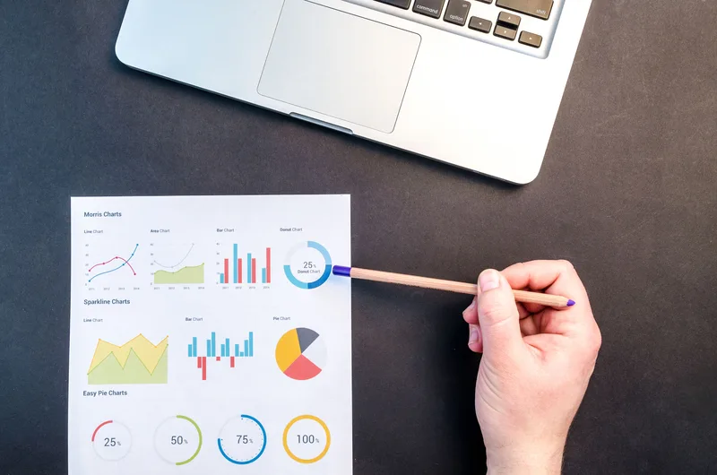 Hand reviewing colorful data charts and graphs on a desk next to a laptop
