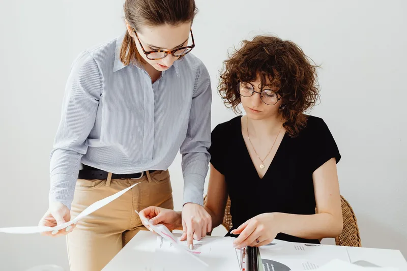 Two businesswomen reviewing documents at a workspace, discussing marketing performance
