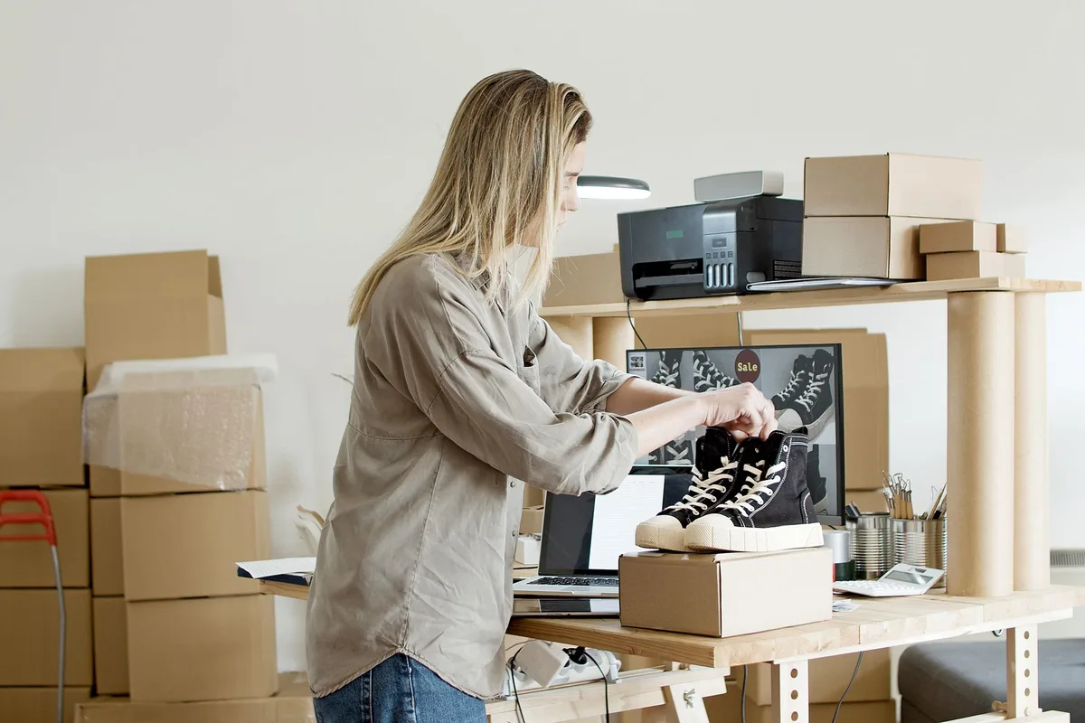 Small ecommerce store owner packing a shoe order at a desk surrounded by shipping boxes, with a product listing open on a laptop
