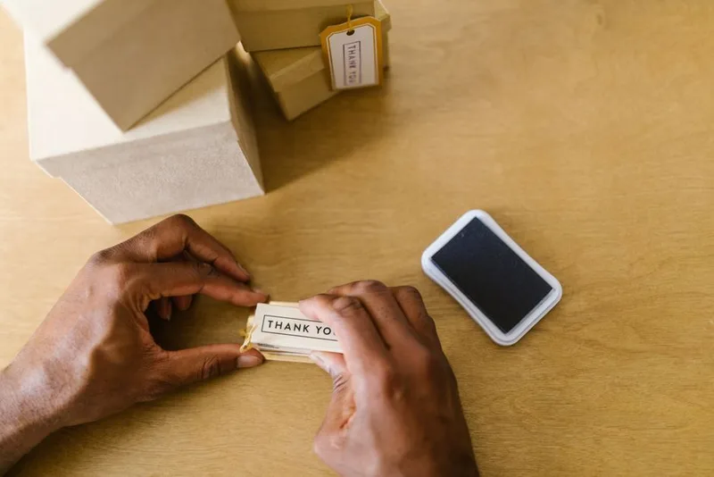 Small business owner attaching a handwritten thank-you tag to a customer’s package next to kraft shipping boxes